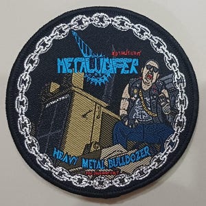 May include: A circular embroidered patch with a black border featuring a silver chain link design. The centre displays a tan bulldozer, a figure in a black vest, and the text "METALUCIFER" and "HEAVY METAL BULLDOZER" in blue, with additional red text.