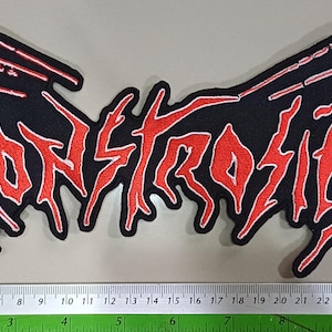 以下が含まれることがあります： 赤色のギザギザした文字で「MONSTROSITY」と書かれた黒いパッチ。パッチの幅は約28cm。テキストは白いステッチで縁取られています。パッチは明るい灰色の表面にあります。
