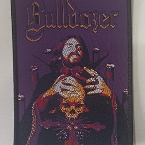 Pode incluir: Patch bordado com fundo roxo e borda preta. O patch apresenta a palavra "Bulldozer" em letras douradas. Uma figura com uma caveira e uma capa escura está no centro. O patch é retangular.