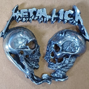 May include: A silver metal pin featuring the word "METALLICA" in a stylized font, with two skull designs on either side of the word.