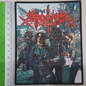 May include: A rectangular embroidered patch featuring a band in dark clothing with a red logo reading Sarcofago at the top. A green ruler is placed along the left side, showing the patch measures approximately 6 inches in height.