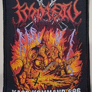 Puede incluir: Un parche rectangular negro con una ilustración detallada y ardiente. El diseño presenta una figura con cuernos con una ametralladora, llamas, relámpagos y el texto "KAOS KOMMAND 696". La palabra "TORMENT" también está presente en rojo.