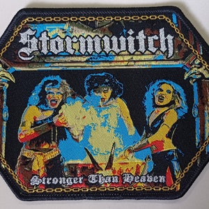 Puede incluir: Un parche bordado negro con un borde de cadena dorada. El parche presenta una banda llamada "Stormwitch" con las palabras "Stronger Than Heaven" debajo del nombre de la banda. La imagen muestra tres figuras frente a un fondo azul y amarillo.