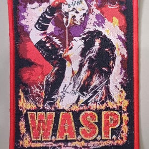 Peut inclure: Écusson brodé avec un graphisme rock and roll. Il représente une figure buvant dans un crâne, des flammes et le nom du groupe "WASP" en lettres rouges. L'écusson a une bordure rouge.