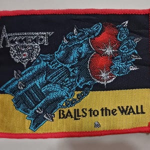 Può includere: Una toppa ricamata nera e rossa con il nome della band "ACCEPT" e le parole "Balls to the Wall".