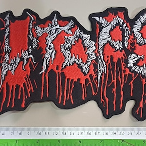May include: Embroidered patch with the word "ATROCITY" in white, stylized lettering over a red background with dripping blood effect. The patch has a black border and is approximately 11 inches long.