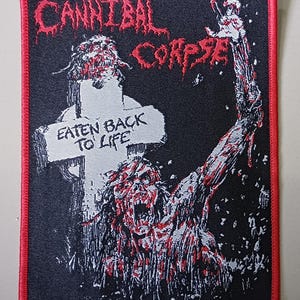 Puede incluir: Parche bordado con las palabras "CANNIBAL CORPSE" en letras rojas y dentadas. El parche presenta un zombi que se levanta de una tumba, con el texto "EATEN BACK TO LIFE" en una lápida. Fondo negro y borde rojo.