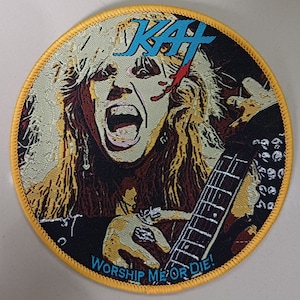 Puede incluir: Un parche redondo, cosido en amarillo, con un fondo negro que presenta un retrato de una persona con cabello largo y rubio, una raya roja en la cara y una guitarra. El texto "KMFDM" está en azul y blanco sobre la cabeza de la persona. El texto "WORSHIP ME OR DIE!" está en azul y blanco en la parte inferior del parche.