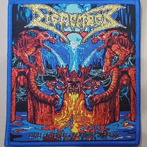 Op de afbeelding: Een vierkante geborduurde patch met een blauwe rand en de bandnaam Dismember in gouden letters bovenaan. De afbeelding toont twee rode skeletachtige wezens op stenen pilaren met een centrale schedel en stromende lava, met de tekst Like An Ever Flowing Stream.