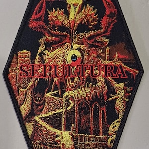 May include: A black and gold embroidered patch featuring the band name "SEPULTURA" and the album title "ARISE" in a gothic font. The patch depicts a detailed image of a demonic figure with a large eye in the centre.