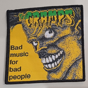 Op de afbeelding: Vierkante patch met een zwarte rand, met een gele achtergrond en een gedetailleerde illustratie van een monstergezicht. De tekst "THE CRAMPS" staat bovenaan in groen en "Bad music for bad people" is verticaal gedrukt.
