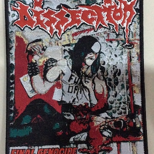 May include: A black and white embroidered patch with a red and white band at the top. The band has the word "Desecration" in white lettering. The patch features a black and white image of a person with long black hair and a white shirt. The person is holding a red object in their right hand. The bottom of the patch has the words "FINAL GENOCIDE" in white lettering.