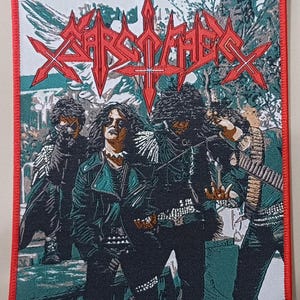 May include: A rectangular embroidered patch with a red border featuring a graphic of four figures in dark clothing and leather jackets. The text Sarcofago is printed in a stylized red font at the top against a green and white outdoor background.