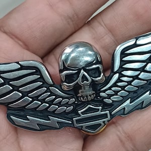 May include: A silver skull with wings, a motorcycle-themed emblem. The skull is centered, with detailed wings extending outwards. The emblem has a dark background with the word "Harley-Davidson" at the bottom.