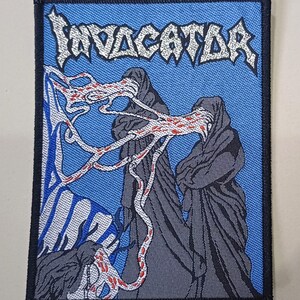 Może przedstawiać: Prostokątna naszywka z napisem "INVACATOR" w stylizowanej czcionce u góry. Naszywka ma niebieskie tło z dwiema postaciami w ciemnych płaszczach i skomplikowanym wzorem białych i czerwonych linii. Naszywka jest obramowana na czarno.