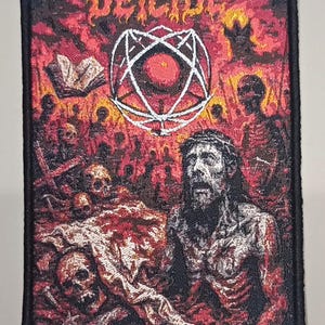 Puede incluir: Parche bordado con la palabra "DEICIDE" en una fuente en forma de llama. El parche presenta un gráfico de una figura con una corona de espinas, rodeada de calaveras y un fondo ardiente. Un borde negro enmarca el diseño.