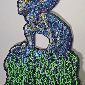May include: An embroidered patch featuring a blue and purple alien figure in a seated, contemplative pose. The alien is positioned above a dense, intricate base of bright neon green, root-like or vine-like structures with a dark black border.