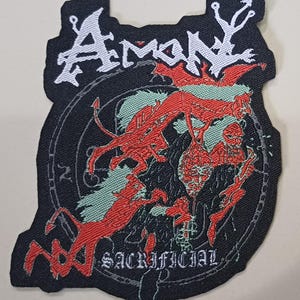 May include: Black patch with the word "Amon" in white gothic lettering. The patch features a red and black illustration of demonic figures and the word "Sacrificial". The patch has a unique, irregular shape.