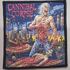 Puede incluir: Un parche cuadrado con las palabras "CANNIBAL CORPSE" en rojo, sobre una escena grotesca de zombis y sangre. El parche tiene un fondo azul y un borde negro.