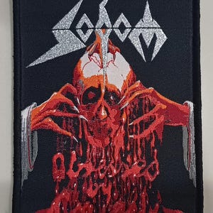 Könnte beinhalten: Schwarzer rechteckiger Aufnäher mit dem Bandnamen "Sodom" in Silber über einer Grafik eines roten Schädels mit tropfendem Blut. Der Schädel hat rote Hände, die sich ausstrecken. Der Aufnäher hat einen schwarzen Rand.