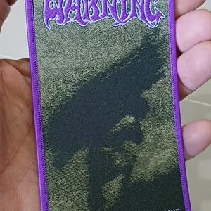 May include: A rectangular patch with a purple border and a textured background. The word "WARNING" is in purple, stylized lettering at the top. Below is a dark, abstract design, and the text "WATCHING FROM A DISTANCE" is at the bottom.