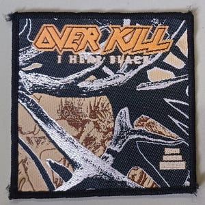 May include: Square patch with the text "OVERKILL" in orange block letters above "I HEAR BLACK" in white. The background features a camouflage pattern in black, tan, and silver. The patch has a black border.