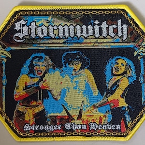 Könnte beinhalten: Ein sechseckiger schwarzer und gelber Patch mit einem silbernen Glitzerband am Rand. Der Patch zeigt ein Band mit dem Text "Stormwitch" in silberner Glitzer Schrift. Unter dem Band befindet sich ein Bild von drei Frauen in schwarzen Lederoutfits mit roten und gelben Akzenten. Die Frauen stehen vor einem blauen und gelben Hintergrund mit einem Kessel in der Mitte. Der Text "Stronger Than Heaven" ist in silberner Glitzer Schrift am unteren Rand des Patches gedruckt.