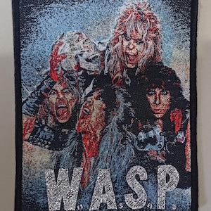 May include: A black and white embroidered patch with a band's logo. The logo features a group of four people with long hair and a silver glitter-like text that reads "W.A.S.P."