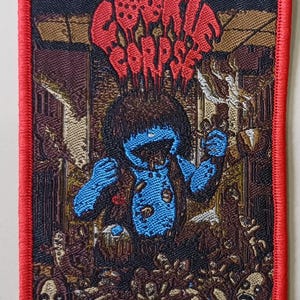 Puede incluir: Parche bordado con borde rojo que presenta un monstruo azul con el texto "COOKIE CORPSE" en rojo. El monstruo está rodeado por un fondo oscuro y detallado con calaveras.