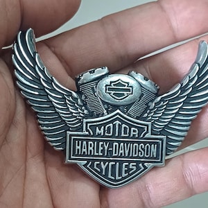 May include: A silver Harley-Davidson emblem with a winged design. The emblem features the words "MOTOR HARLEY-DAVIDSON CYCLES" in a rectangular frame, with engine details at the centre.