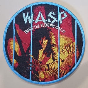 May include: Circular patch with a blue trim, displaying the band name "W.A.S.P." in light blue. The design features a red and yellow background with an image of a person and the text "INSIDE THE ELECTRIC CIRCUS".