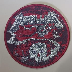 Puede incluir: Parche redondo bordado con fondo rojo y negro. El parche presenta el nombre de la banda "Metallica" en plata, junto con un gráfico de calavera. El parche tiene un borde rojo.