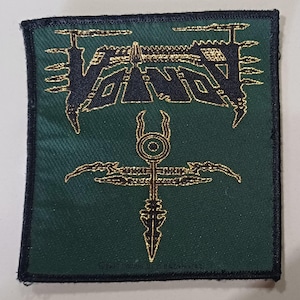 May include: A square fabric patch with a dark green background and a black border. The patch features the word VOIVOD in a stylized black font with gold metallic outlines, positioned above a gold metallic symbol resembling a spear or cross.