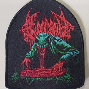 May include: Embroidered patch with a black background and a gothic arch shape. The patch features red, stylized text at the top and a green and blue creature with red details. The creature appears to be vomiting red liquid.