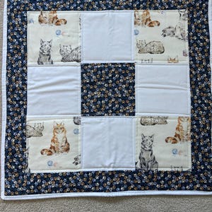 May include: A handmade quilt with a patchwork design. The quilt has a central square of white fabric surrounded by squares with cat illustrations. The border is a dark blue fabric with a paw print pattern. The quilt is approximately 91 cm square.