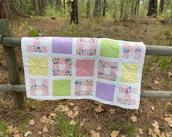 Puede incluir: Un edredón hecho a mano con diseño de patchwork. Presenta cuadrados en colores pastel: rosa, amarillo, verde y lavanda, con patrones florales y de punto de cruz. El edredón está sobre una valla de madera en un entorno natural.