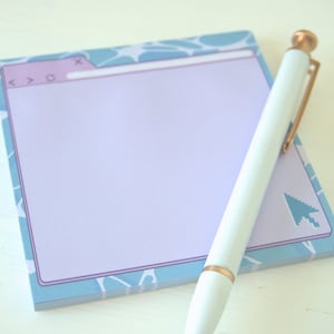 May include: A notepad with a light purple interior and a blue and white patterned border, designed to resemble a browser window. A white pen with gold accents rests on the notepad, next to a pixelated arrow icon.
