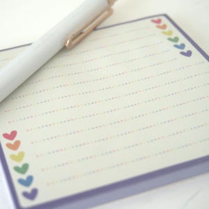 May include: A white pen with gold accents rests on a lined notepad. The notepad has a purple border and is decorated with colorful heart-shaped designs. The notepad is cream-colored with dotted lines.