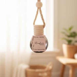 May include: A hanging car air freshener with a clear glass bottle and a wooden top. The label reads "Rose Creek Moonflower Nectar Car Freshener." The freshener is suspended by a natural rope and wooden bead.
