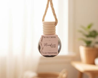 Puede incluir: Un pequeño frasco de vidrio redondo para ambientador de coche con tapa de madera clara y cuerda beige para colgar. La botella tiene una etiqueta rectangular con el texto Rose Creek Moonflower Nectar Car Freshener y un dibujo floral.