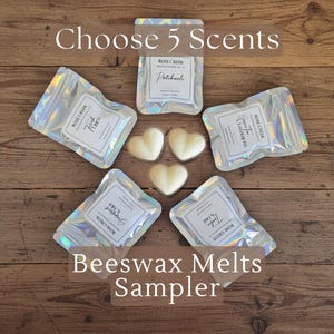 Op de afbeelding: Een bovenaanzicht van een houten oppervlak met vijf iriserende zakjes en drie hartvormige wassmelten. De zakjes zijn voorzien van het label "Rose Creek" en verschillende geurnamen. De tekst "Choose 5 Scents" en "Beeswax Melts Sampler" is ook zichtbaar.