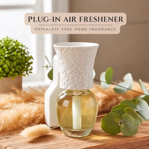 May include: A plug-in air freshener with a white, floral-embossed ceramic cover and a clear glass base containing golden liquid. The text PLUG-IN AIR FRESHENER and PHTHALATE-FREE HOME FRAGRANCE is displayed in a tan banner above the device.