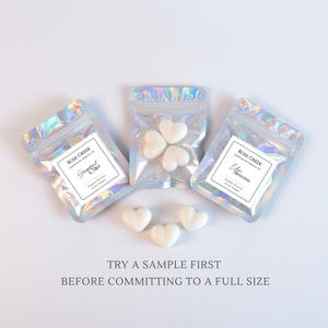 Op de afbeelding: Drie iriserende cellofaan zakjes met hartvormige wax melts. Elk zakje heeft een label met de tekst "Rose Creek" en geurnamen. Losse hartvormige melts zijn verspreid rond de zakjes. De tekst "TRY A SAMPLE FIRST BEFORE COMMITTING TO A FULL SIZE" staat onderaan.