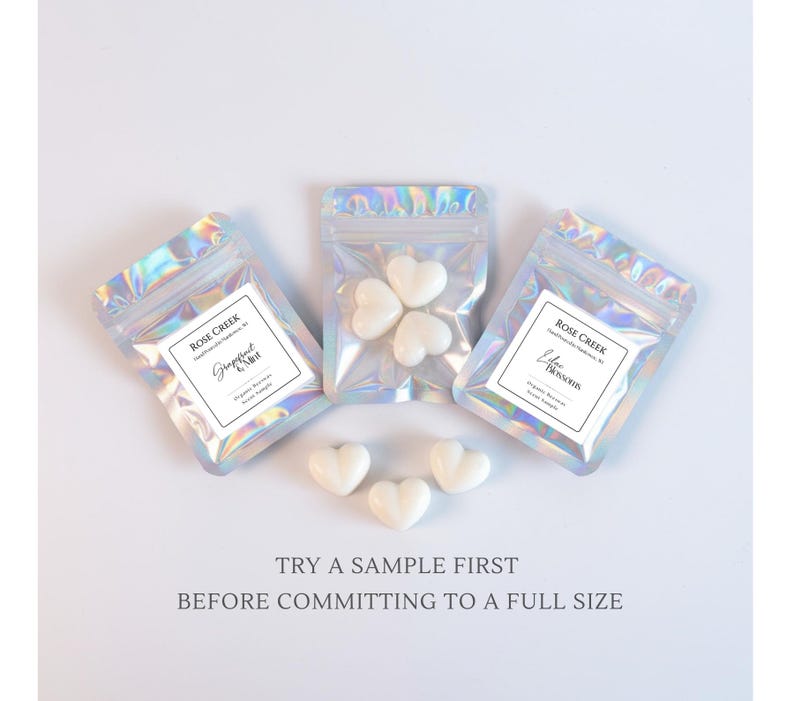 Op de afbeelding: Drie iriserende cellofaan zakjes met hartvormige wax melts. Elk zakje heeft een label met de tekst "Rose Creek" en geurnamen. Losse hartvormige melts zijn verspreid rond de zakjes. De tekst "TRY A SAMPLE FIRST BEFORE COMMITTING TO A FULL SIZE" staat onderaan.
