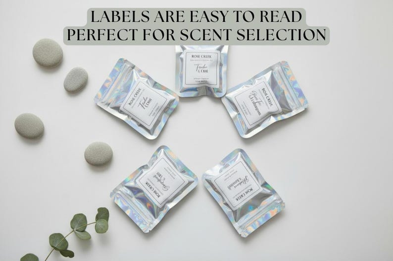 Op de afbeelding: Vijf zilveren zakjes met witte etiketten, gerangschikt in een cirkelvormig patroon. De etiketten lezen "Rose Creek" en geurnamen. De tekst "LABELS ARE EASY TO READ PERFECT FOR SCENT SELECTION" staat bovenaan. Grijze stenen en groen zijn ook aanwezig.