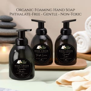Op de afbeelding: Drie donker amberkleurige plastic flessen Rose Creek schuimende handzeep met zwarte pompdoseerders. Elke fles heeft een rond zwart etiket met een witte calla lelie illustratie en tekst waaronder Cucumber Melon, Fresh Linen en Coconut Lime.