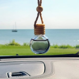 May include: A clear glass car air freshener with a wooden top hanging from a brown string. The air freshener is hanging from the rearview mirror of a car. The car is parked in front of a body of water.