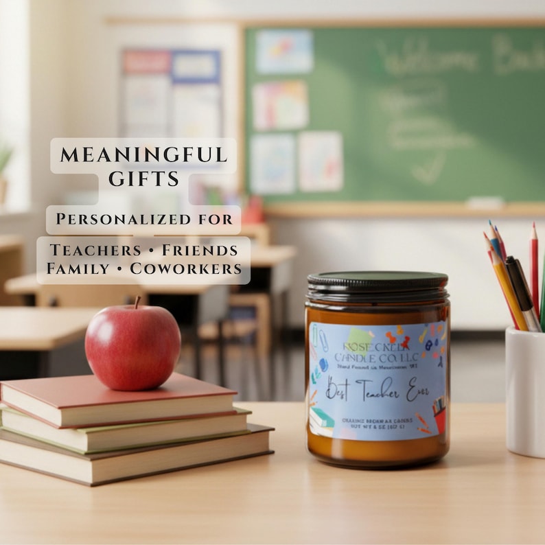 May include: A brown glass candle jar with a label reading Rose Creek Candle Co. LLC and Best Teacher Ever, placed next to a stack of three books topped with a red apple and a white pencil holder containing colored pencils and pens.