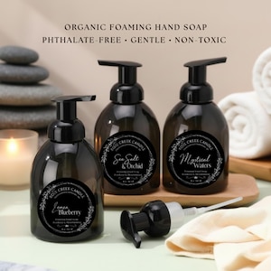 May include: Three dark brown foaming hand soap bottles with black pumps, labeled "Lemon Blueberry", "Sea Salt & Orchid", and "Mystical Waters". The text "Organic Foaming Hand Soap" and "Phthalate-Free • Gentle • Non-Toxic" are at the top. A fourth pump is lying on its side.