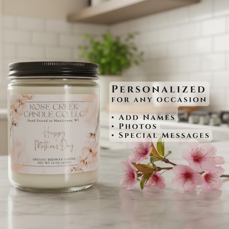 May include: A 16 oz organic beeswax candle in a clear glass jar with a black lid. The label features floral illustrations, the text Rose Creek Candle Co. LLC, Happy Mother's Day, and net weight 16 oz (454 g). A branch of pink cherry blossoms sits nearby.